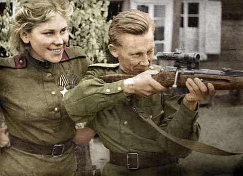 Photo of a smiling Roza Shanina looking over the shoulder of a male comrade while he holds her rifle and looks down the barrel