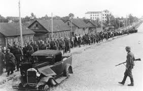 Photo of Soviet soldier standing in the middle of a street with a rifle, next to a broken down car, watching hundreds of Soviet soldiers walk in a line down the side of the street